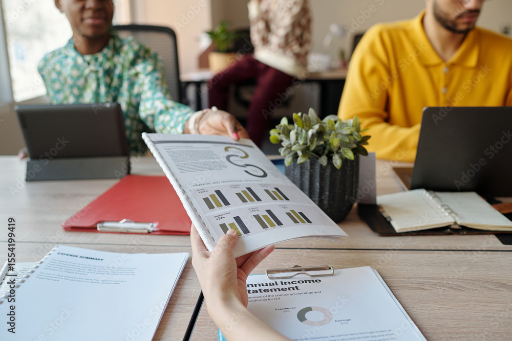 Poster woman handing business report to black colleague while sitting at coworking table with young adult m