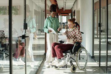 Young adult Black woman standing and discussing documents with Caucasian man sitting in wheelchair...