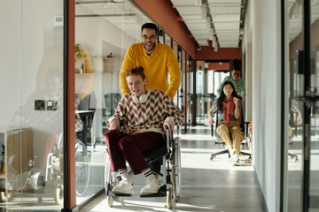 Young Black man pushing Caucasian man with red hair in wheelchair through modern office, young female colleagues having fun in background