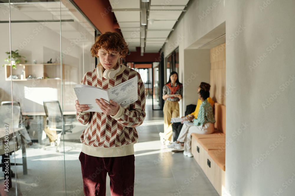 Poster caucasian man with curly red hair standing in modern coworking space reading documents while two you
