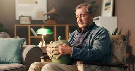 Senior, sad man and home with urn or memory for grief, loss or mourning in thought on chair....