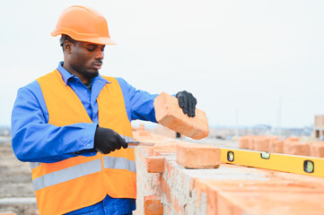An African American construction worker, in work clothes and a hard hat, is building a brick house