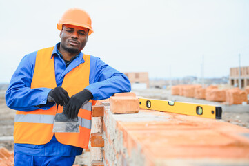 An African American construction worker, in work clothes and a hard hat, is building a brick house