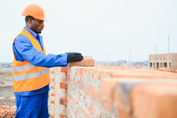 An African American construction worker, in work clothes and a hard hat, is building a brick house