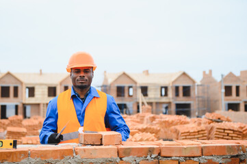 An African American construction worker, in work clothes and a hard hat, is building a brick house