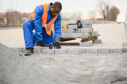 African american master in gloves lays paving stones in layers. Garden brick pathway paving by professional paver worker. Laying gray concrete paving slabs in house courtyard on sand foundation base - Powered by Adobe