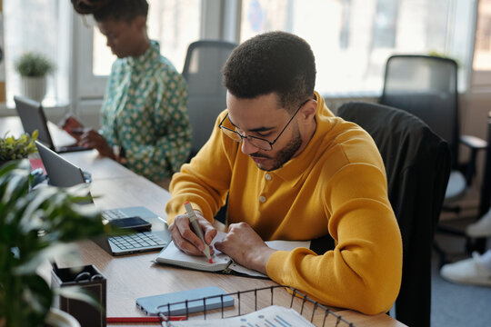 Young Black man wearing yellow sweater writing in notebook at shared desk while young adult Black woman in patterned shirt working on laptop