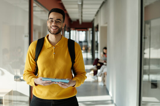 Biracial man with short hair and beard wearing yellow sweater and glasses standing in coworking space holding digital tablet and smiling at camera - Powered by Adobe