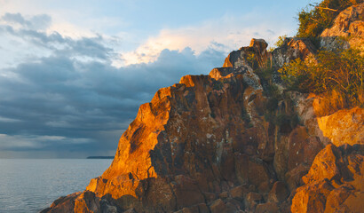 Rocky coastline glows under the evening light beside a calm sea and cloudy sky