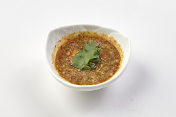 Bowl of spicy dipping sauce garnished with fresh cilantro leaf, featuring vibrant mix of herbs, chili, and spices, served in white ceramic dish on plain white background