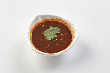 Small white bowl filled with spicy red chili sauce garnished with fresh cilantro leaf, placed on clean white background. sauce appears rich and flavorful, with visible chili flakes and herbs