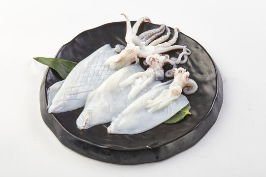 Fresh raw squid arranged on black ceramic plate with green leaves, showcasing its tentacles and body parts, ready for cooking or seafood preparation