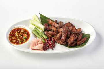 Crispy sun dried pork served on white plate with fresh cucumber slices, pickled ginger, dried chili, and spicy dipping sauce garnished with green onions, creating flavorful Thai dish