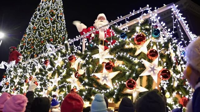 Santa Claus waves cheerfully from a festive float adorned with lights and decorations during a lively Christmas parade. A large, sparkling tree stands tall in the background