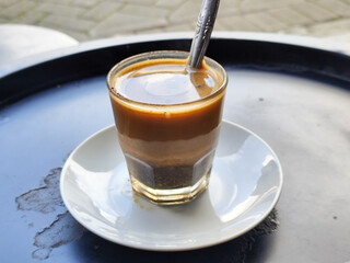 This close-up photo shows a cup of hot black coffee served on a small white plate with a spoon inside, placed on a black table in a roadside stall.