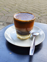 This close-up photo shows a cup of hot black coffee served on a small white plate with a spoon inside, placed on a black table in a roadside stall.