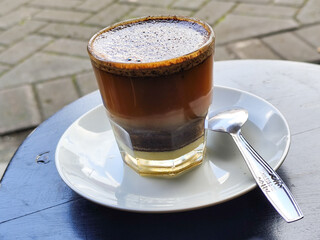 This close-up photo shows a cup of hot black coffee served on a small white plate with a spoon inside, placed on a black table in a roadside stall.