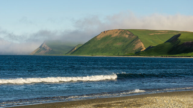 Scenic coastal landscape featuring rolling green hills, gentle waves lapping at the sandy shore, and misty mountains in the background, creating a serene natural environment