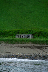 Abandoned wooden cabin sits on sandy shore, surrounded by lush green grass and steep hillside,...