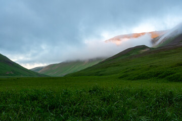 Lush green valley surrounded by majestic mountains, with soft clouds drifting over peaks, creating...