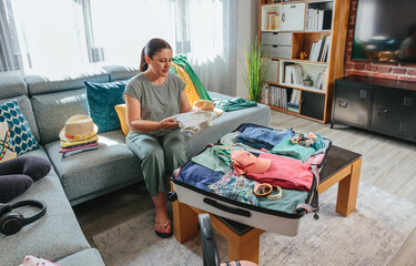 Portrait of happy woman packing clothes in suitcase on table in living room with clothes and...