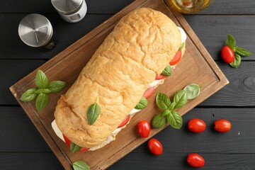 Tasty sandwich with mozzarella cheese, tomatoes, basil and spices on black wooden table, flat lay