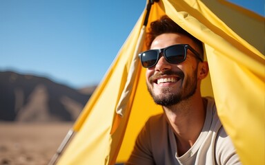 Happy young brunette bearded man tourist in sunglasses peeping out of touristic tent, smiling looking into distance, enjoying vacation adventure in camping on sandy mountains and blue sky background