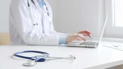 Closeup of blue stethoscope is lying on the table near female doctor working on laptop, highlighting digital tools transforming healthcare diagnostics and patient documentation. Medicine concept