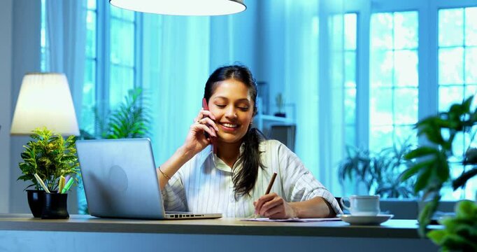 Businesswoman taking notes on phone call sitting at computer desk in modern cozy office, Indian Asian female using pencil to jot down pointers while talking on phone and working on laptop