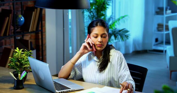 Businesswoman taking notes on phone call sitting at computer desk in modern cozy office, Indian Asian female using pencil to jot down pointers while talking on phone and working on laptop