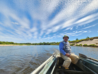 Fototapeta premium Man is sitting in a boat on a river. The sky is blue and there are clouds. fisherman