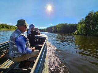 Man and a woman are sitting in a boat on a lake. The man is wearing a striped shirt and a hat