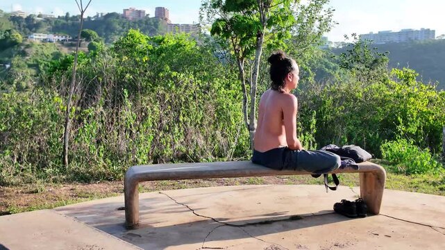 Shirtless man with hair in a bun sits on bench, cross-legged on top of hill, green landscape with distant buildings. South America
