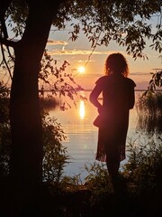 Woman silhouette is standing in front of a tree by a lake. The sun is setting and the water is...