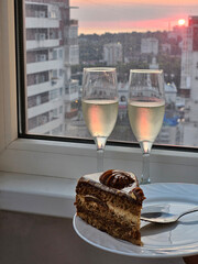 Slice of cake is on a plate in front of two champagne glasses. The cake is cut in half and placed on a white plate near sunset city ocean Sochi