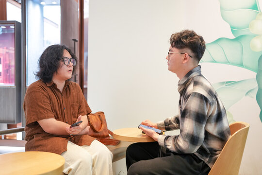 Two Chinese men in their early twenties, both wearing glasses, are sitting together in a stylish cafe, casually talking and laughing. It is a warm May afternoon in Huangpu District, Shanghai, China.