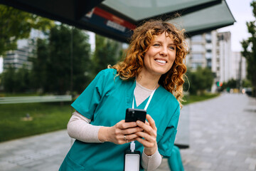 Healthcare worker smiles while checking phone at a bus stop during a sunny day