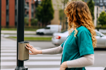 Woman in medical scrubs pushes crosswalk button at busy urban intersection on a sunny day