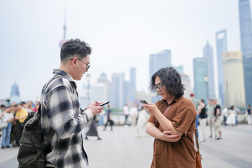 Fototapeta premium Two young Chinese men in their early twenties, both wearing glasses, face each other and talk along the riverside with tall skyscrapers in view across the water in May 2025 in Shanghai, China.