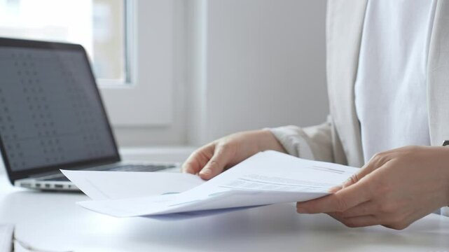 Professional female analyzing financial reports, reviewing charts on laptop and paper documents while seated at desk in corporate workspace. Business people concept