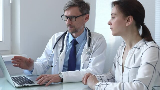Male doctor showing medical test results on a laptop to a female patient, discussing diagnosis and treatment options in a bright medical office. Medicine and health care