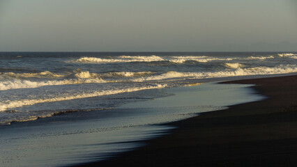 Serene beach landscape at sunset, with gentle waves lapping against dark sand, reflecting warm hues of the sky, creating a tranquil atmosphere for relaxation and contemplation