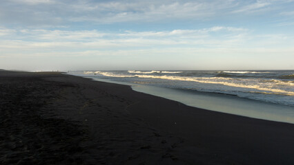 Serene beach landscape at dusk, with gentle waves lapping against dark sand, under a sky filled...
