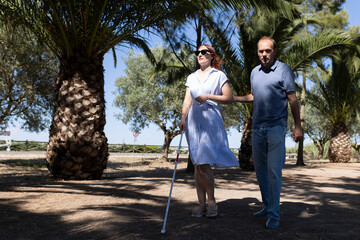 Man guiding blind woman with white cane in park with palm trees