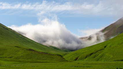 Lush green valley surrounded by majestic mountains, with soft clouds drifting through the...