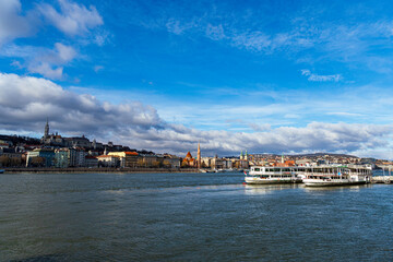 Obraz premium Panoramic view of the Danube River with historic buildings and boats in Budapest, Hungary under a vibrant blue sky.