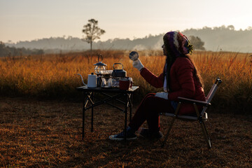 Fototapeta premium woman sitting outdoors with camping gear, including a table, chair, and a steam lamp. She is enjoying her morning coffee in a serene, natural setting.
