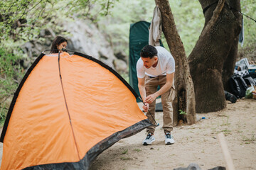 A man and a woman collaborate setting up an orange tent in a forested area, creating a cozy camping...