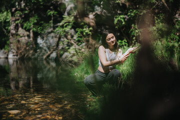 A woman peacefully interacts with her surroundings while kneeling near a calm forest creek.