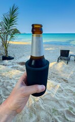 A low-angle, close-up shot captures a hand holding a chilled beer bottle in a black cooler sleeve against the idyllic backdrop of a sun-drenched tropical beach. The golden sand, sparkling turquoise